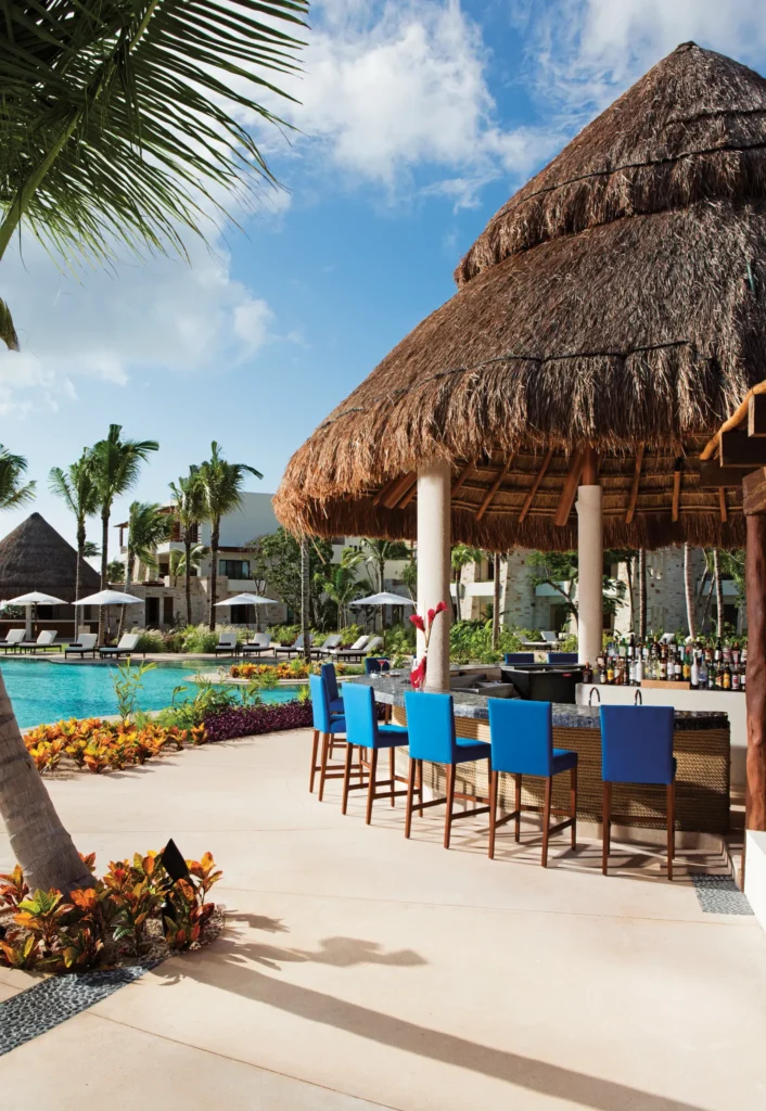 Sugar Reef Poolside Bar at Secrets Akumal Riviera Maya featuring a thatched-roof palapa bar with blue barstools, tropical landscaping, and a sparkling resort pool backdrop.