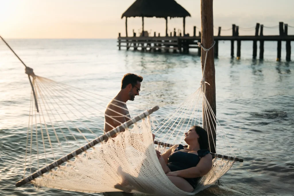 Couple relaxing together in a hammock over the water at Secrets Aura Cozumel during a golden sunset.