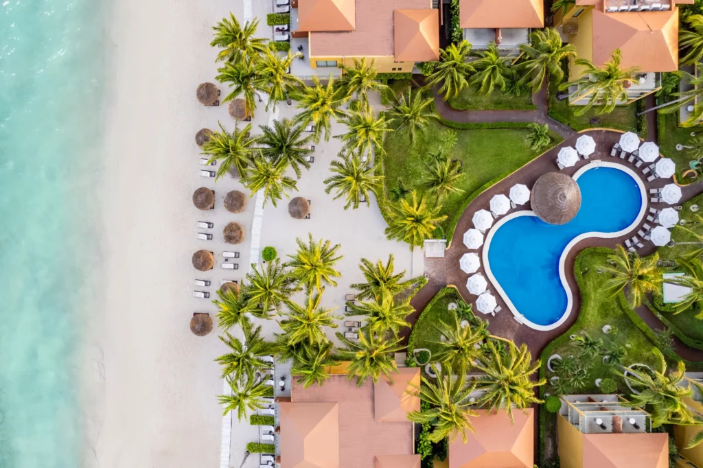 Aerial view of Secrets Aura Cozumel showing beachfront, palm trees, lounge chairs, and curved pool design along the Caribbean Sea.