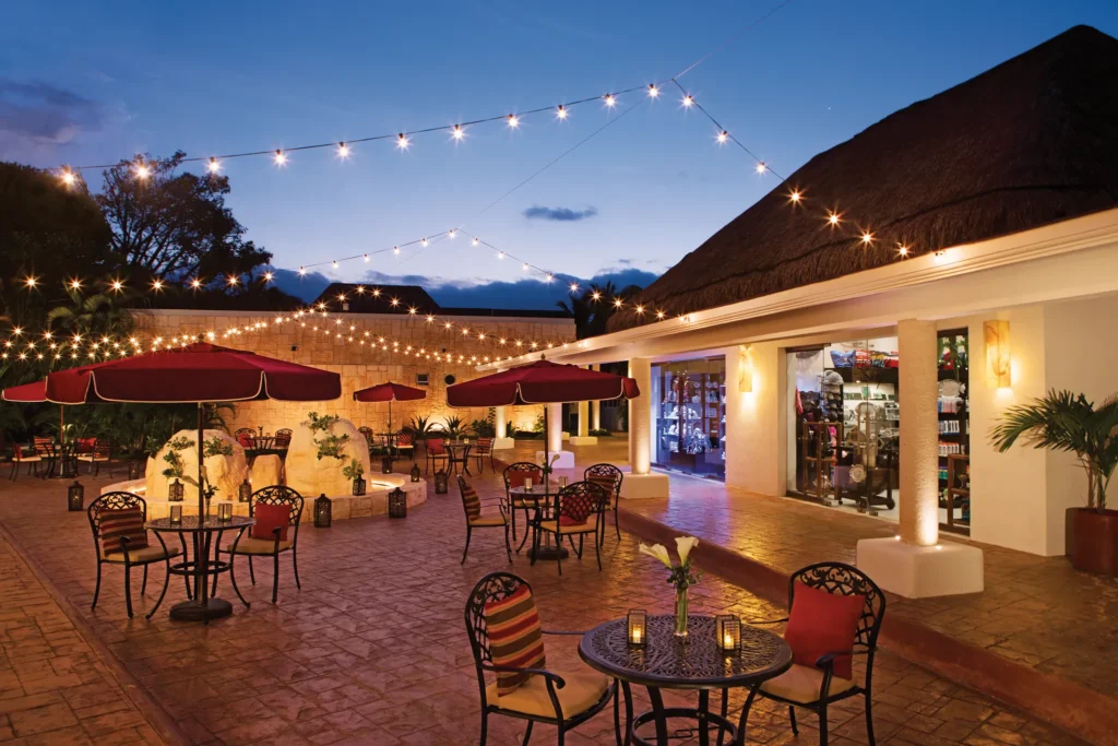 Outdoor plaza at Secrets Aura Cozumel at night with string lights, tables, and umbrellas.