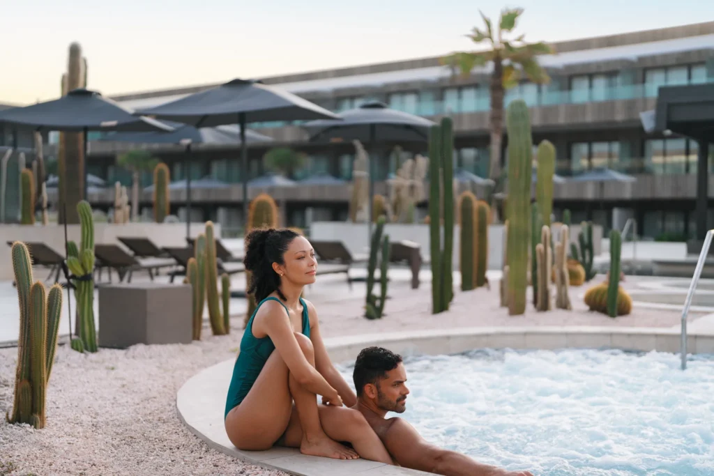 Couple relaxing together in a bubbling hot tub at Secrets Baby Beach Aruba surrounded by modern desert landscaping.
