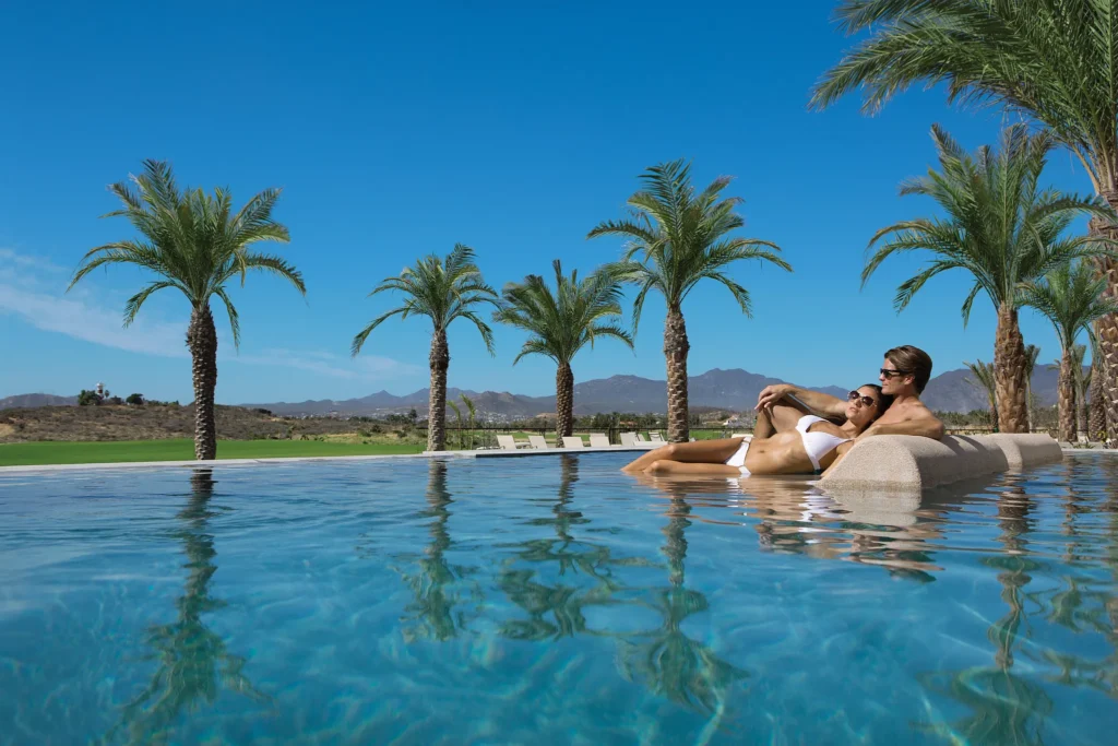 Couple lounging on in-pool beds surrounded by palm trees at Secrets Puerto Los Cabos resort.