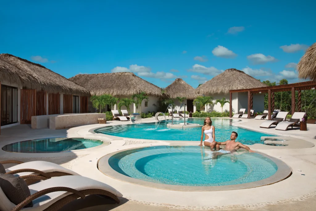 Guests relaxing in the outdoor hydrotherapy circuit pool at Secrets Cap Cana Resort & Spa, surrounded by tropical thatched-roof spa villas in Punta Cana.