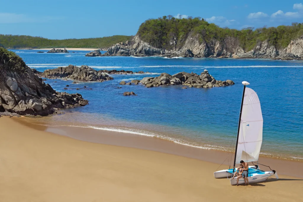 Couple preparing to sail a Hobie Cat along the secluded bay at Secrets Huatulco Resort & Spa, surrounded by turquoise waters and rocky coastline.