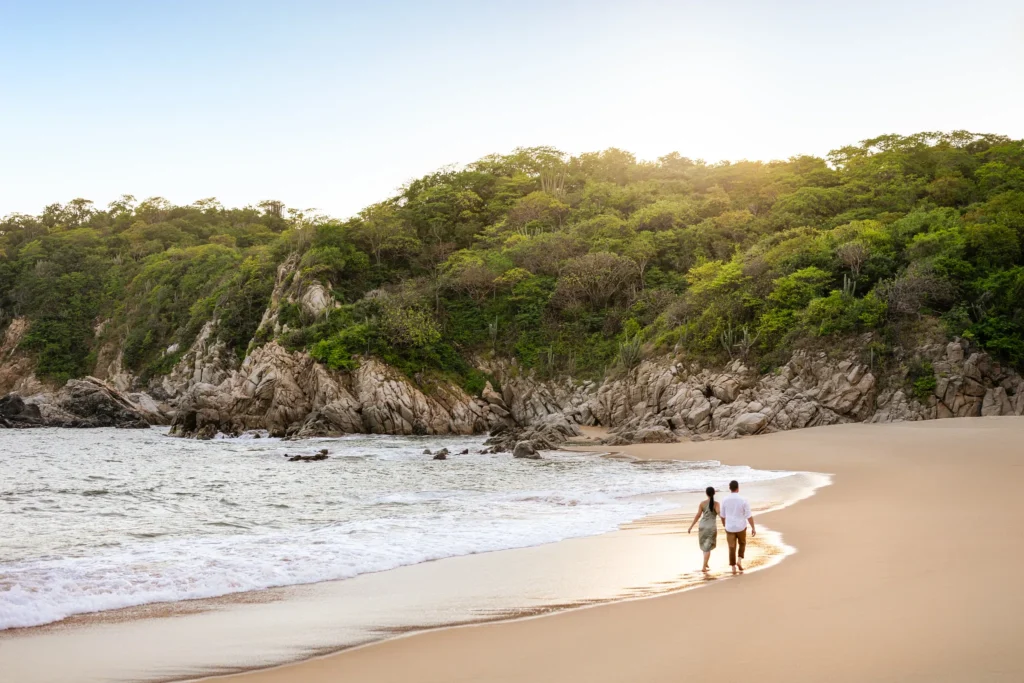 Couple enjoying a serene walk on a secluded beach at Secrets Huatulco Resort & Spa with rocky cliffs and tropical greenery.