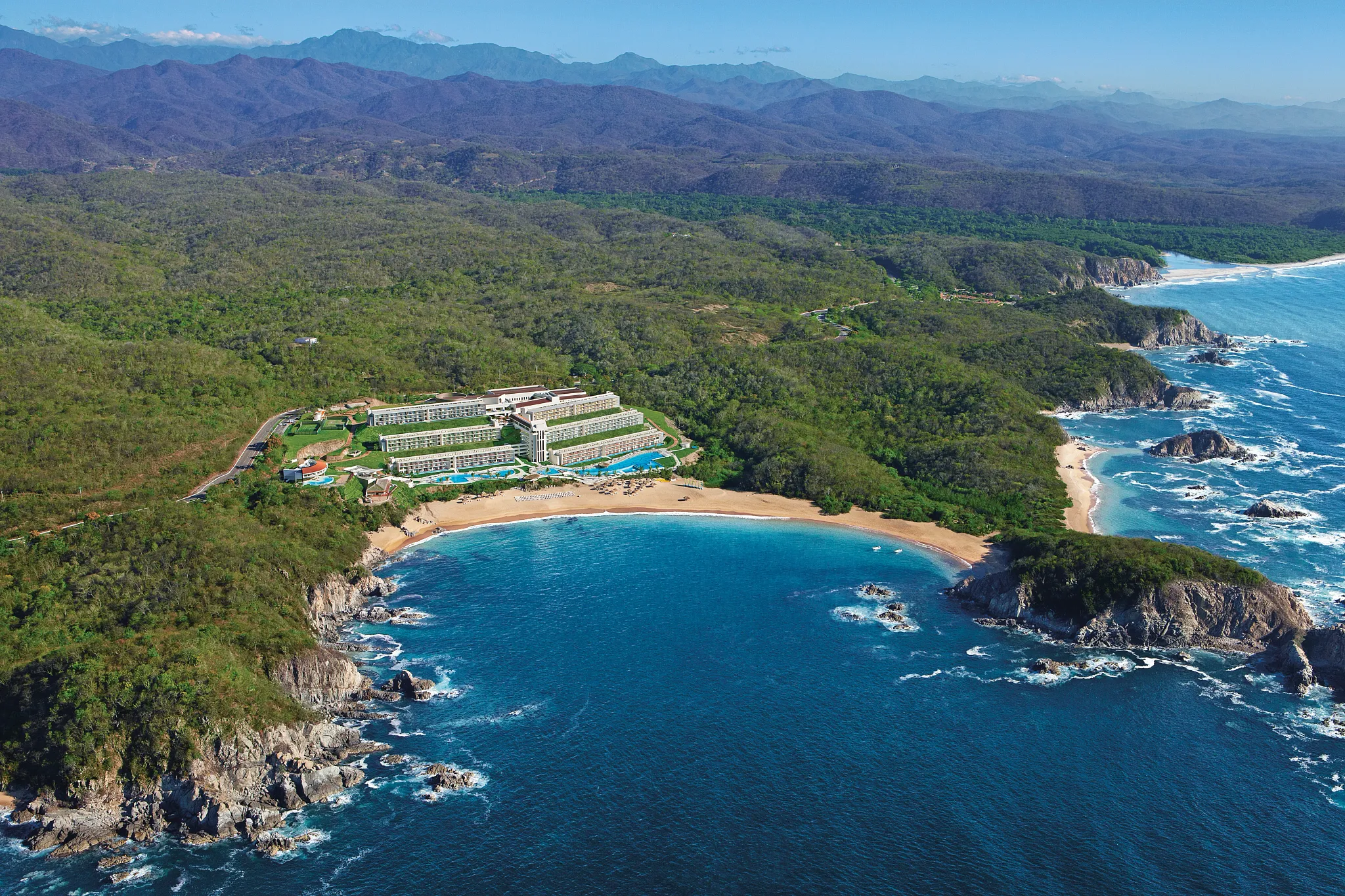 AAerial view of Secrets Huatulco Resort & Spa nestled between lush mountains and a secluded bay along the Pacific coast of Mexico.