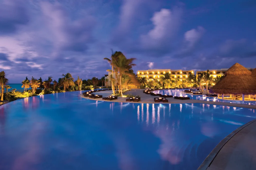 Illuminated infinity pool at night at Secrets Maroma Beach Riviera Cancun with palm trees and resort lights reflecting on the water.