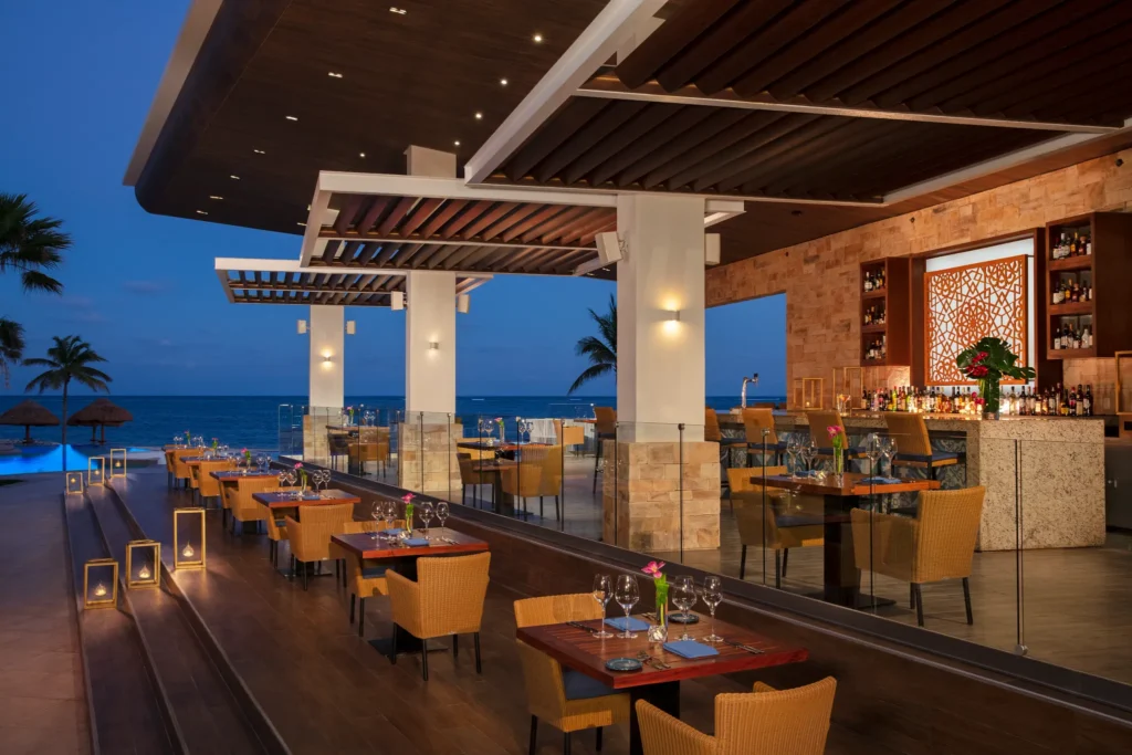 Rosewater restaurant at Secrets Riviera Cancun featuring open-air oceanfront dining, elegant table settings, and a stylish bar terrace at sunset.
