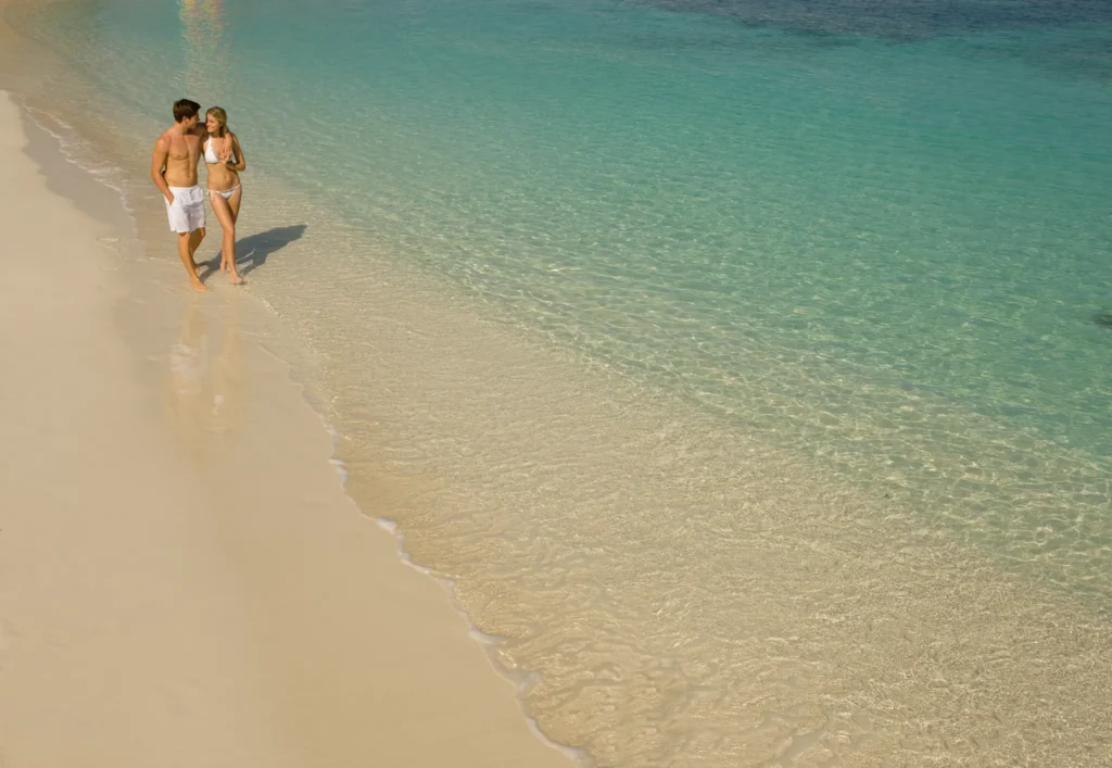 Couple walking along a white-sand beach with clear turquoise water at Secrets St. James Montego Bay, perfect for romantic getaways.