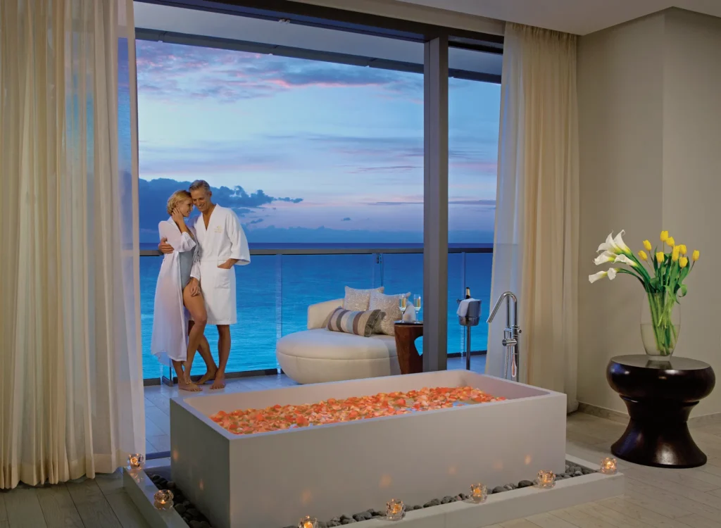 Couple on oceanfront balcony of Preferred Club Honeymoon Suite at Secrets The Vine Cancun with in-room soaking tub and sunset view.