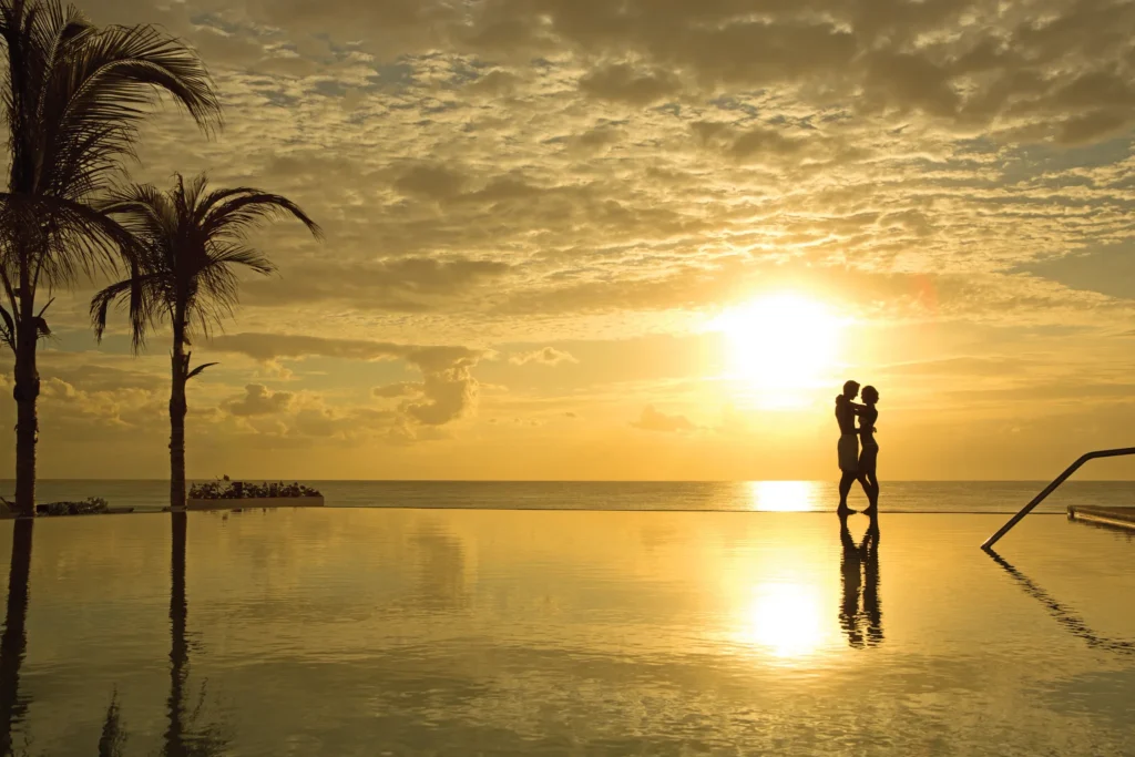 Silhouette of a couple at sunrise by the infinity pool at Secrets The Vine Cancun with golden reflections over the ocean.