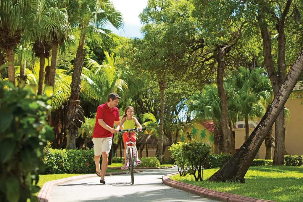 Father helping daughter ride a bike through tropical gardens at Sunscape Curaçao Resort, Spa & Casino.