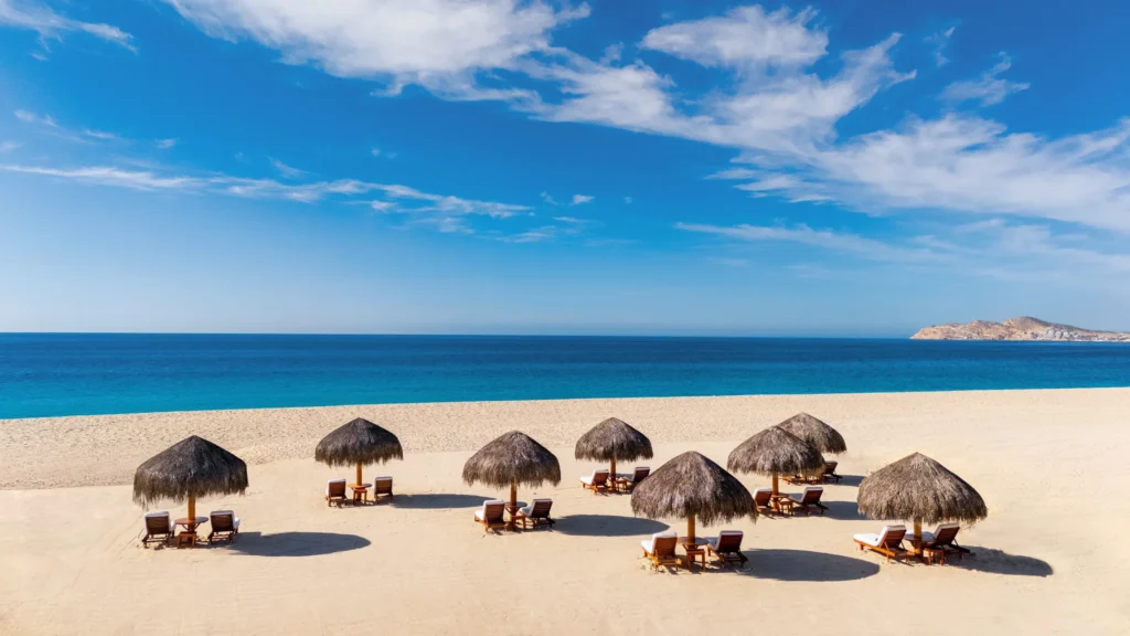 Thatched palapas and lounge chairs on sandy beach at Zoëtry Casa del Mar Los Cabos with ocean backdrop.