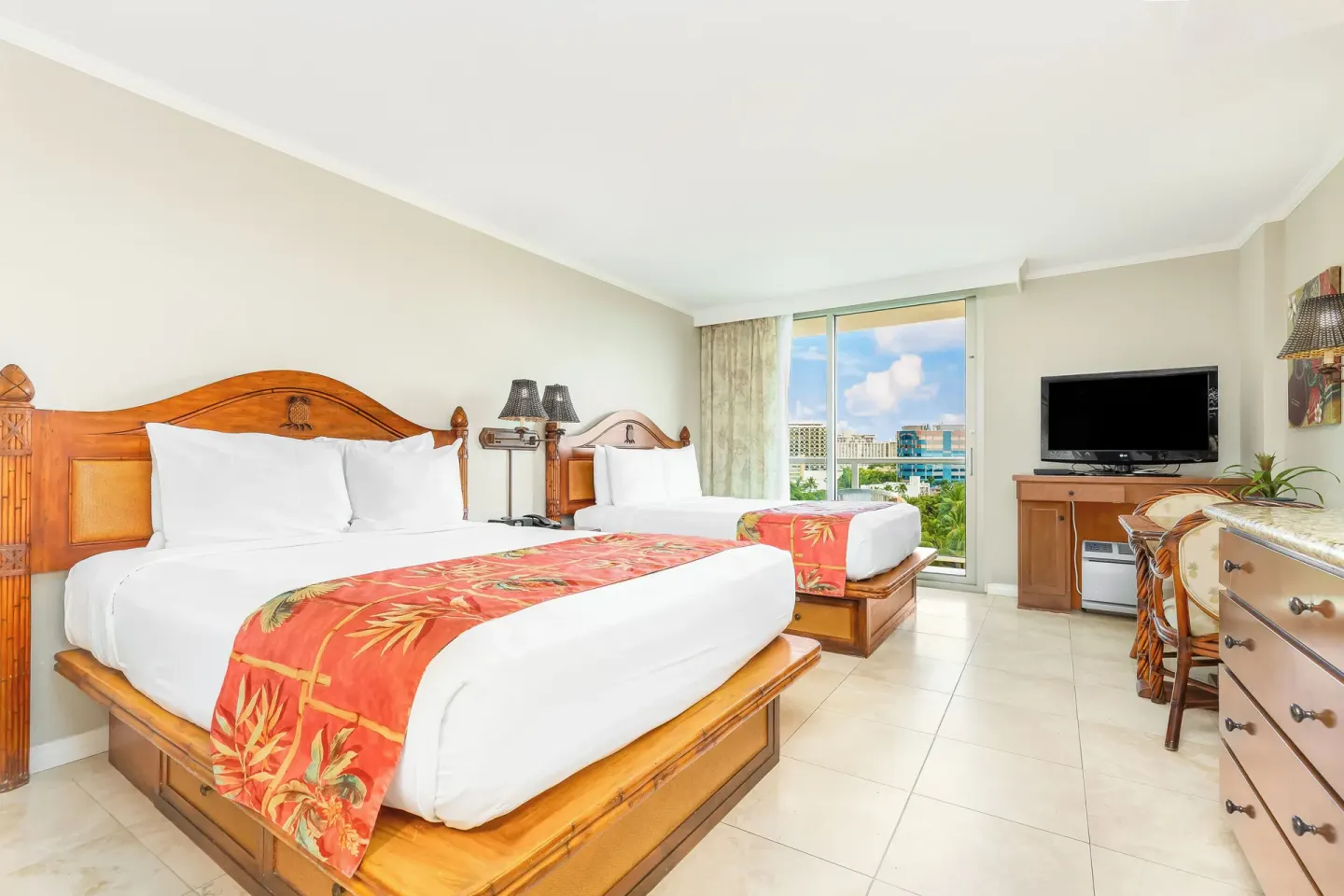 Two beds, a TV, and balcony doors are shown in a guest room at Luana Waikiki Hotel & Suites.