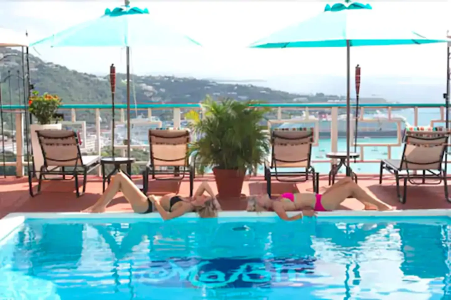 Guests sunbathing by the pool at Mafolie Hotel with turquoise umbrellas and harbor views behind them.