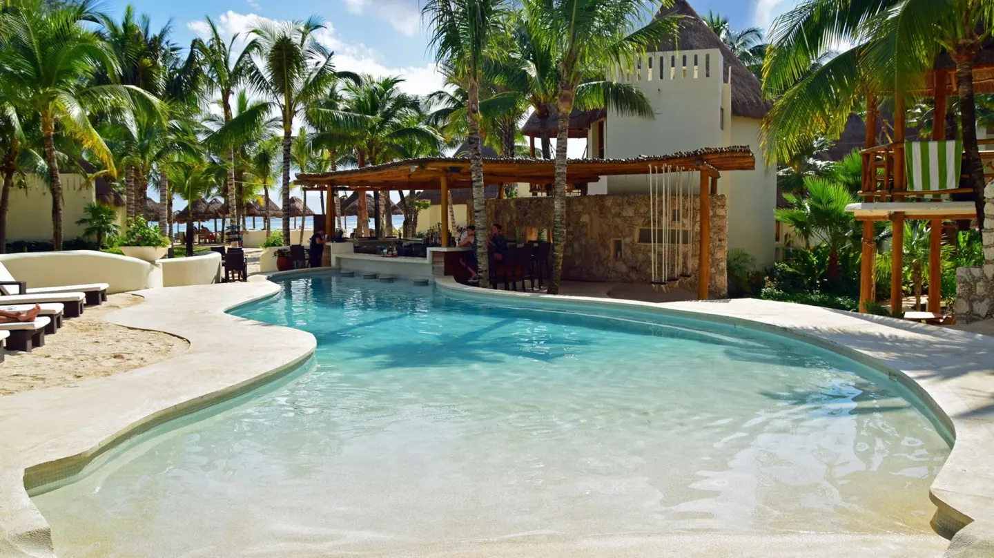 Swim-up pool bar with palms and thatched buildings at Mahekal Beach Front Resort & Spa.