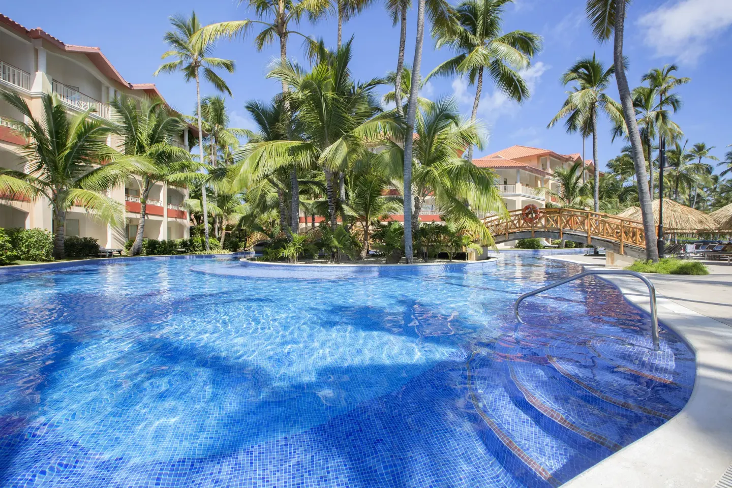 Blue resort pool with palm trees and a wooden bridge at Majestic Elegance Punta Cana.