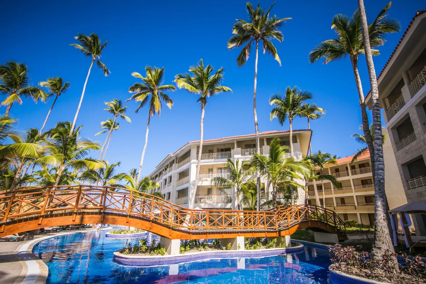 Wooden footbridge over a blue pool beside resort buildings at Majestic Mirage Punta Cana.