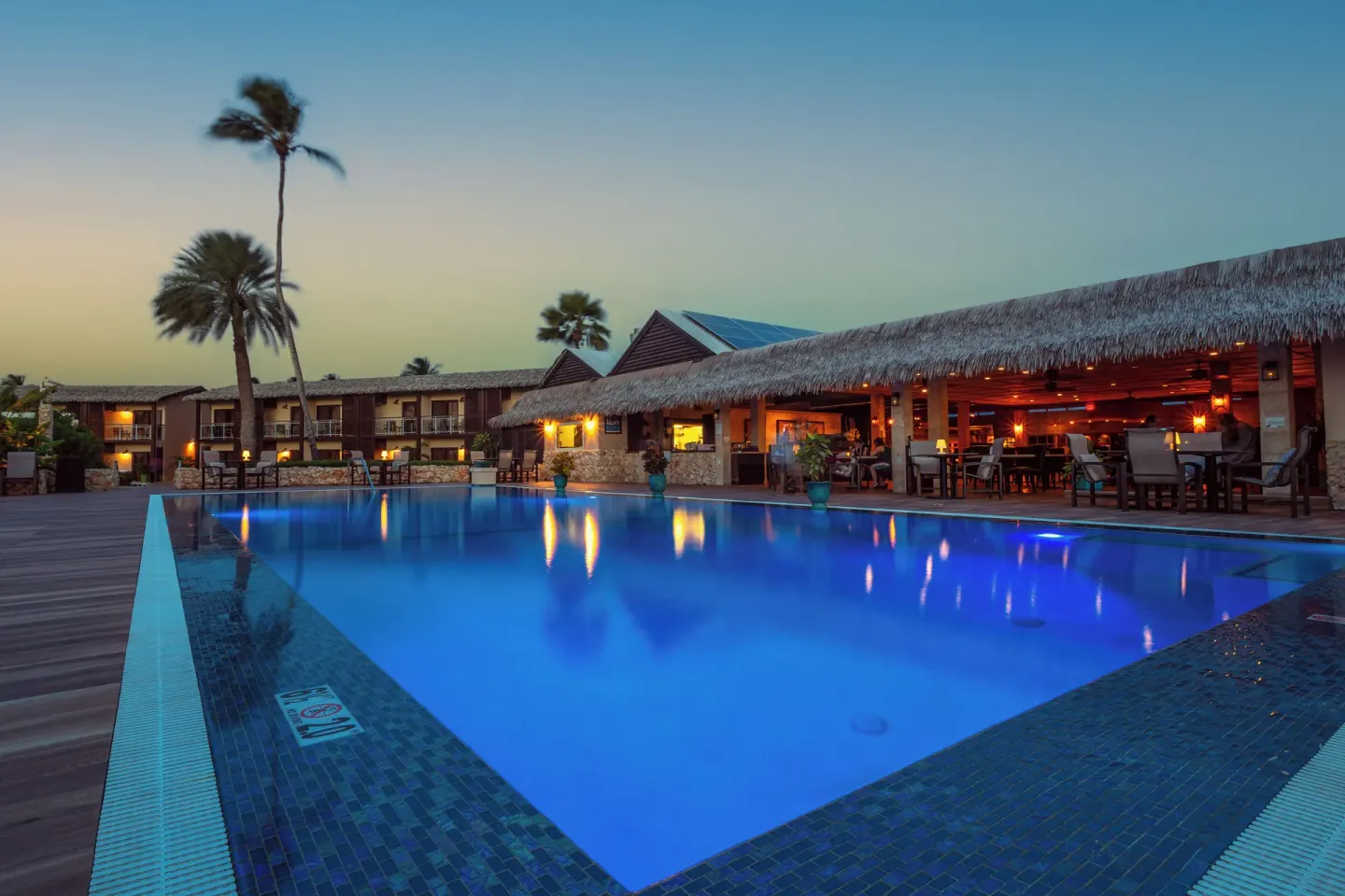 Illuminated pool and thatched dining terrace at Manchebo Beach Resort and Spa at dusk.