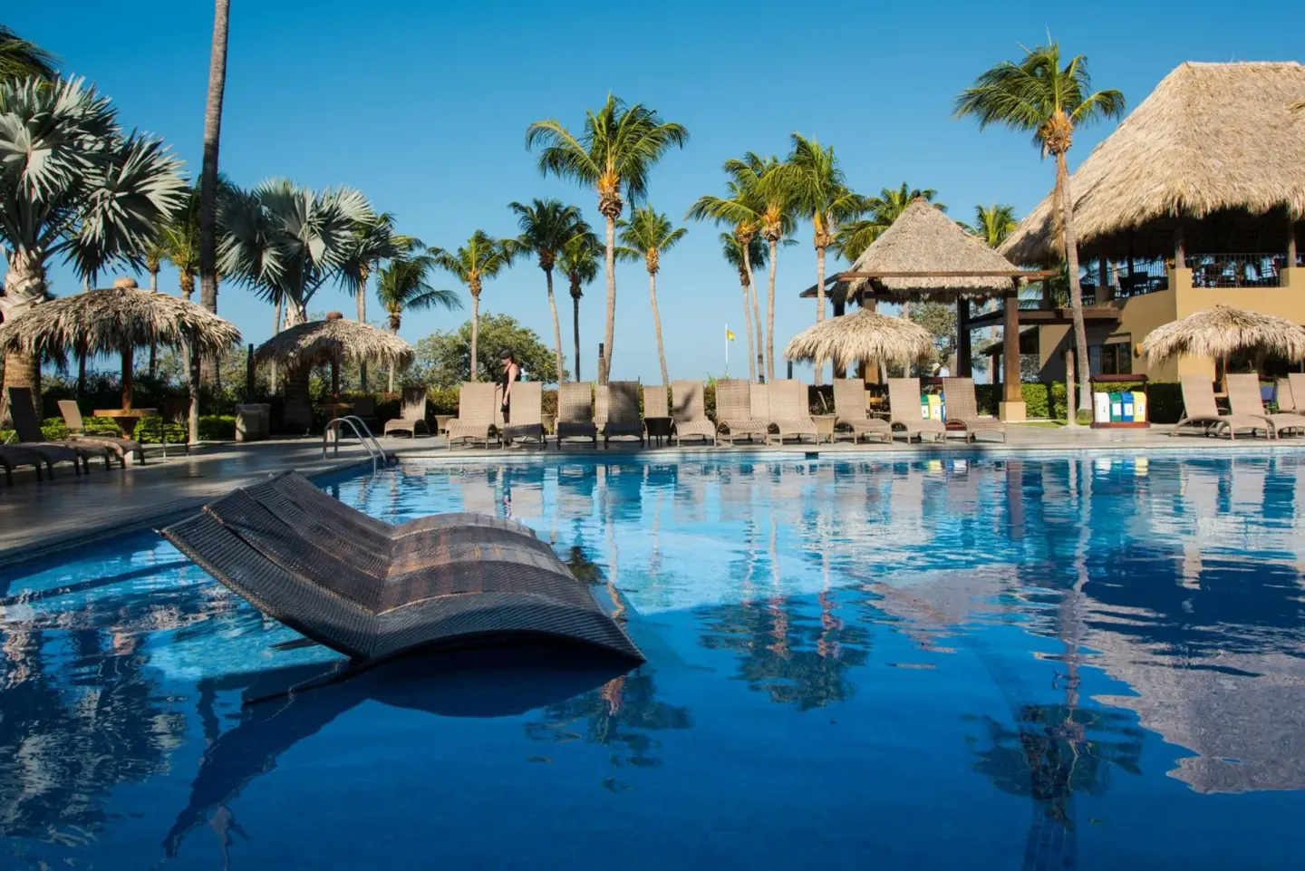 Pool with in-water lounger, palms and thatched shade at Margaritaville Beach Resort Playa Flamingo.