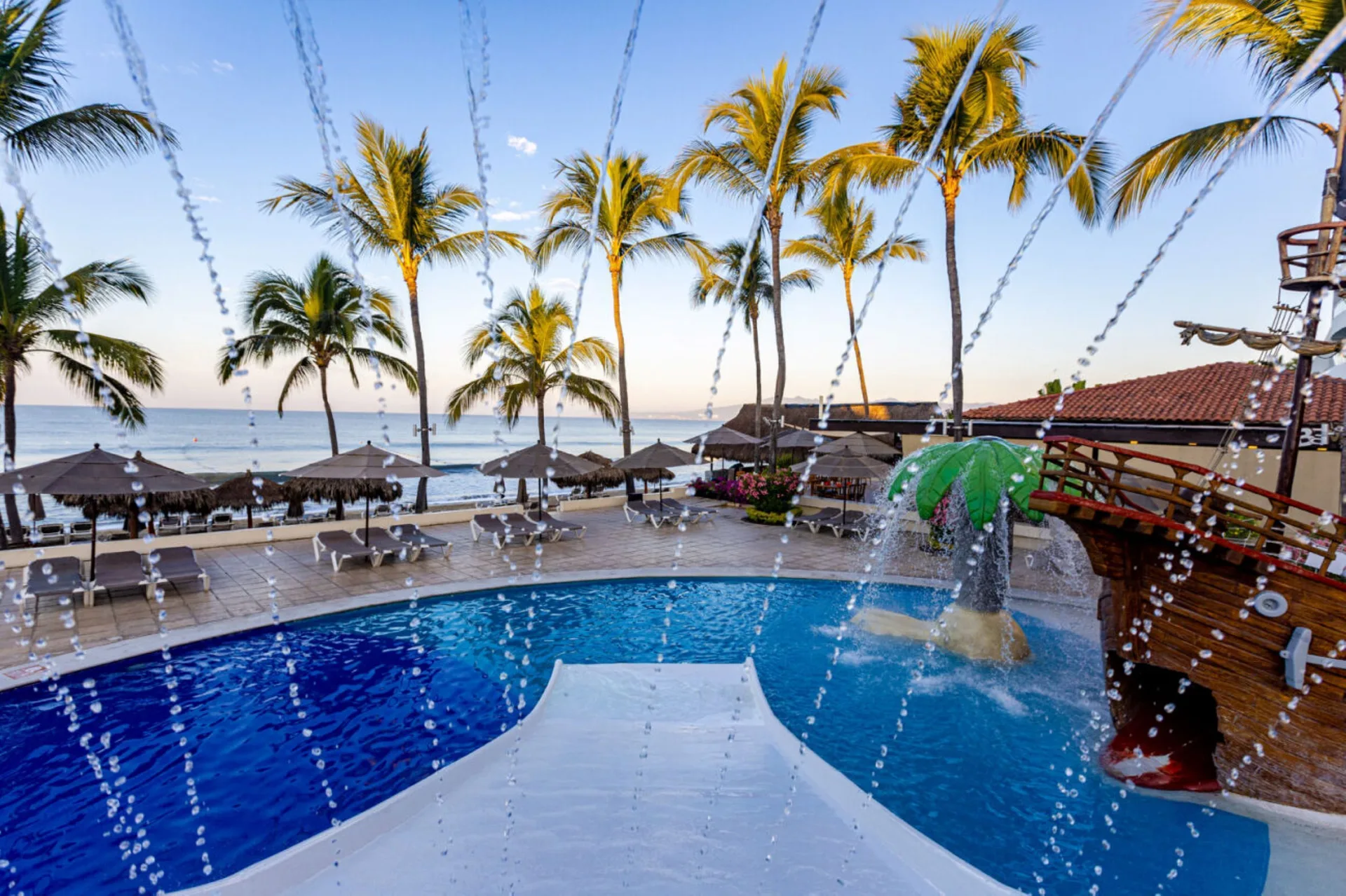 Children's pool slide framed by splash jets and ocean palms at Marival Emotions Resort.