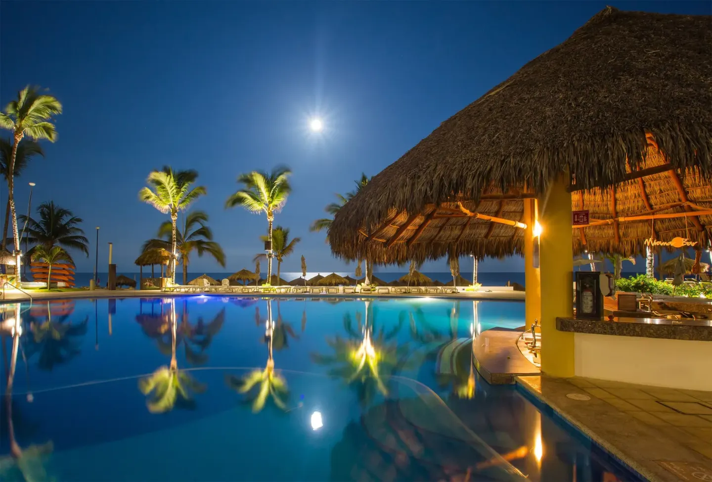Nighttime pool with lit palms, ocean views, and a thatched bar at Marival Emotions Resort.