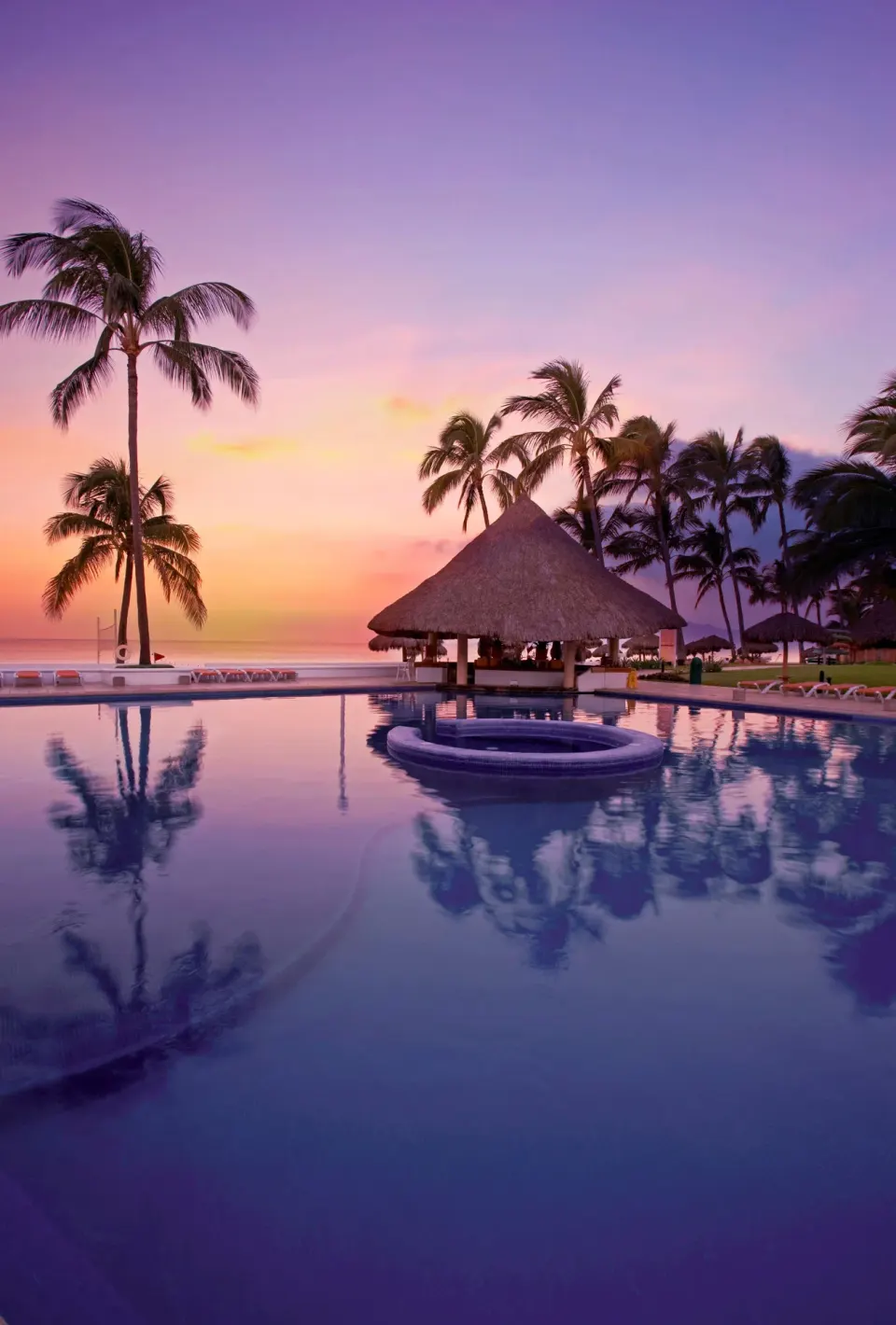 Sunset over the pool with palm trees and a thatched bar at Marival Emotions Resort.
