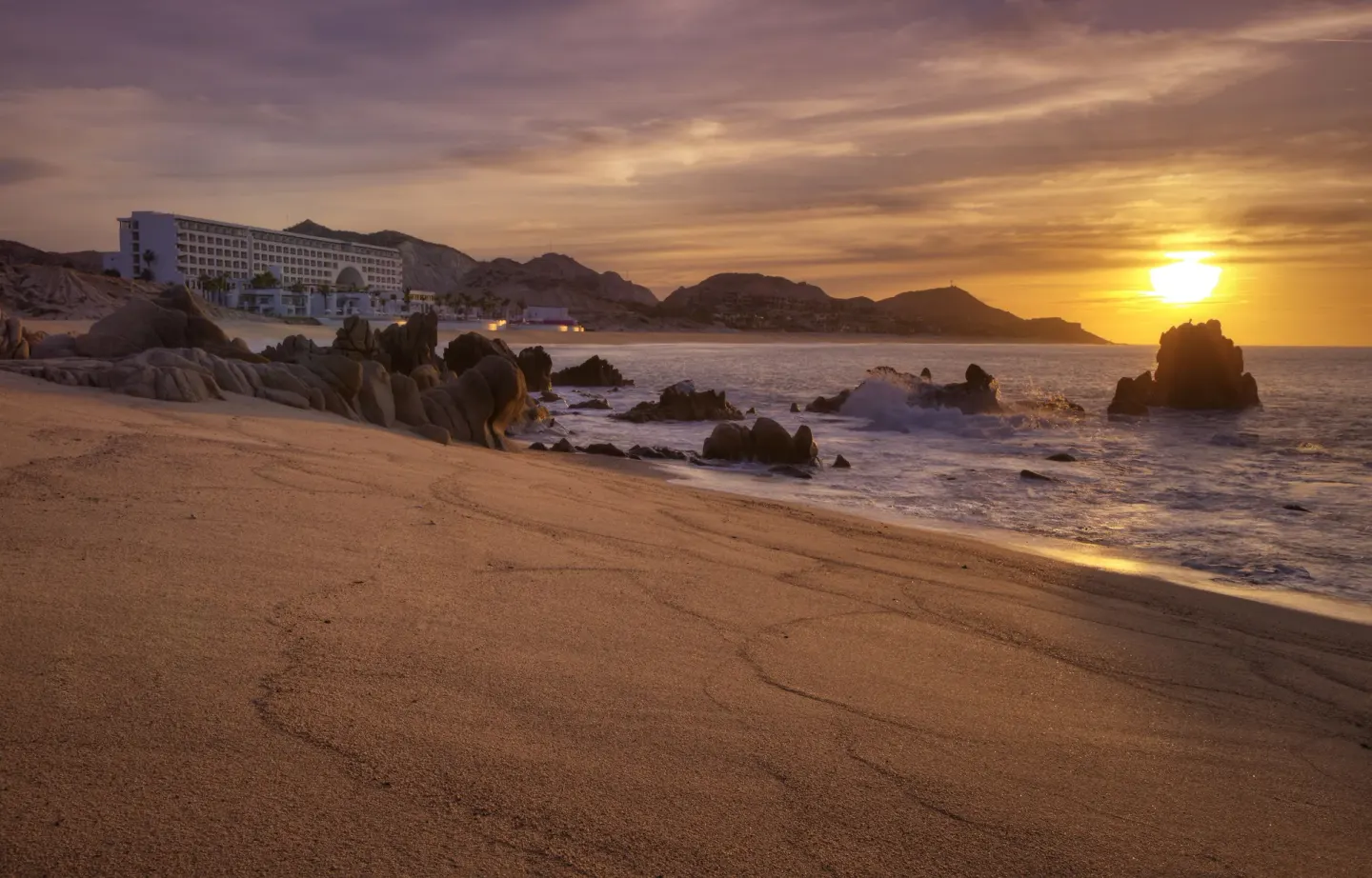 Sunset beach scene with waves, rocks, and the resort building at Marquis Los Cabos.