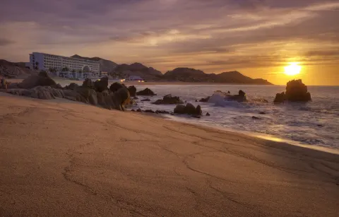 Sunset beach scene with waves, rocks, and the resort building at Marquis Los Cabos.