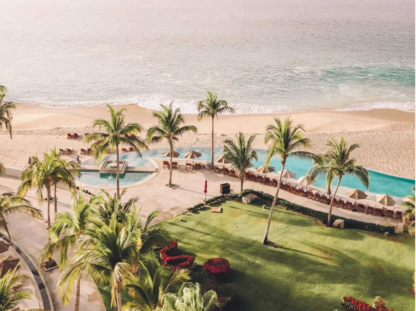 Oceanfront pools, palms, lawn, and beach viewed from above at Marquis Los Cabos.