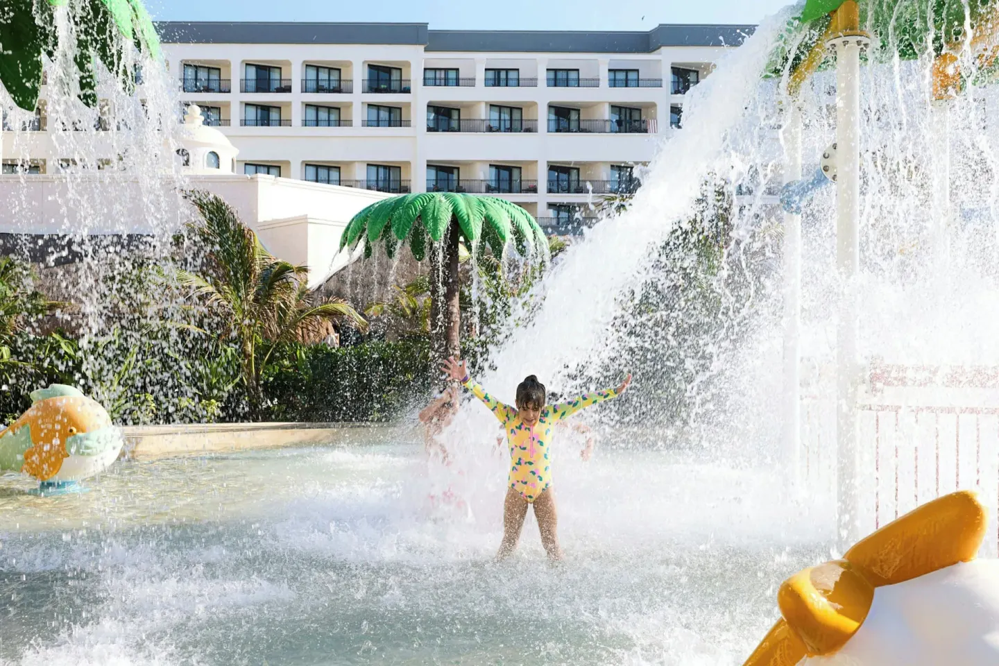 Child stands under a splash bucket in the water play area at Marriott Cancun.