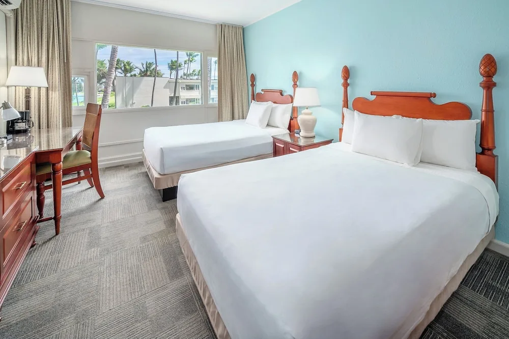Two-bed guest room with desk, blue accent wall, and palm views at Maui Beach Hotel.