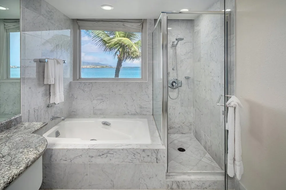 Marble guest bathroom with tub, glass shower, and ocean-view window at Maui Beach Hotel.