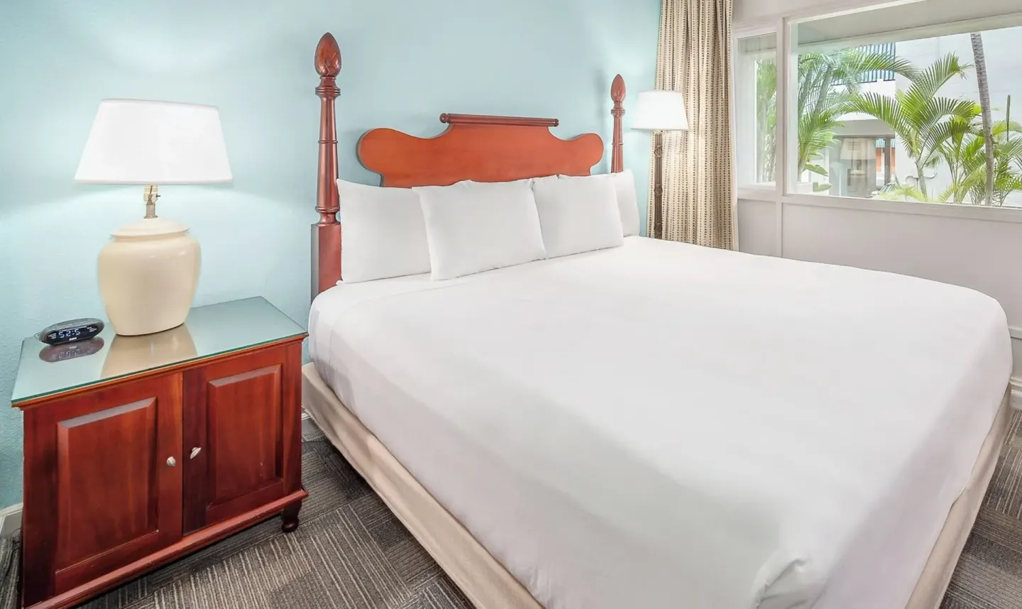 Standard Room One Bed at Maui Beach Hotel with white bedding, wood headboard, nightstand, lamp, and window.