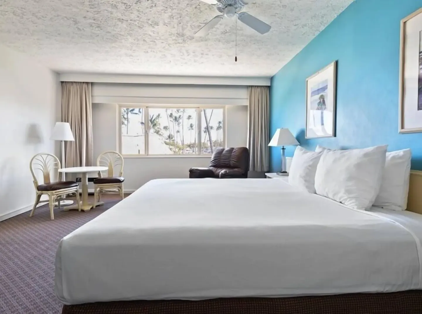 Guest room with king bed, blue accent wall, table, chairs, and lounge chair at Maui Seaside Hotel.