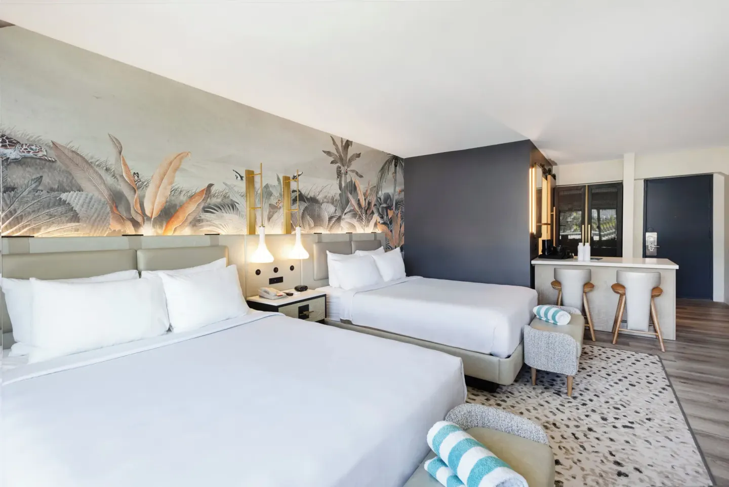 Two-bed guest room with tropical mural, stools, and vanity area at Maui Seaside Hotel.