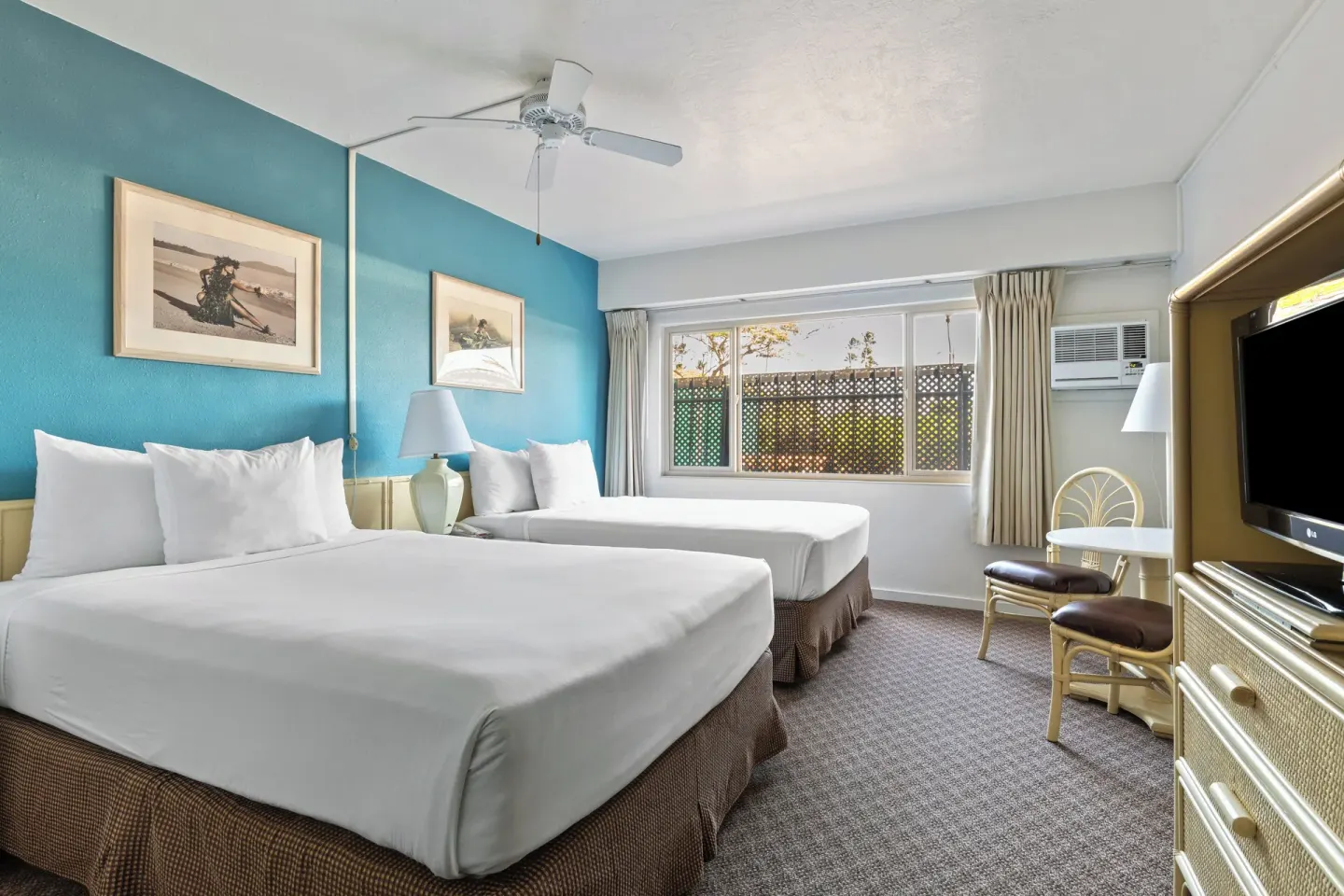 Two-bed guest room with turquoise wall, window, desk, and TV at Maui Seaside Hotel.