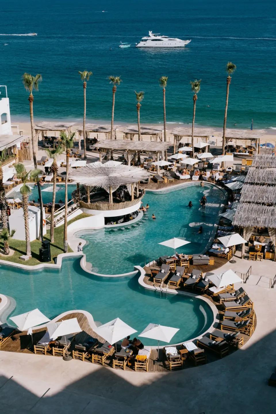 Aerial view of ME Cabo pools, beach seating, palm trees, and the sea.