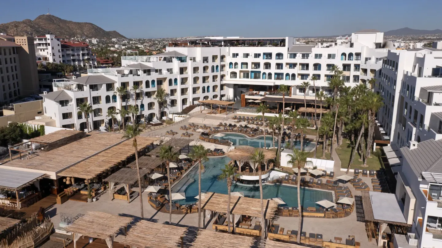 An aerial view shows the pools, palms, loungers, and white resort buildings at ME Cabo.