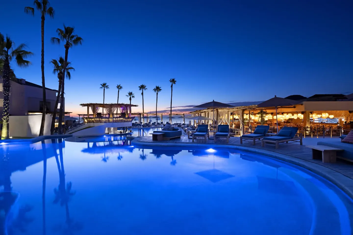 Blue-lit resort pool with palm trees and open-air seating at ME Cabo.
