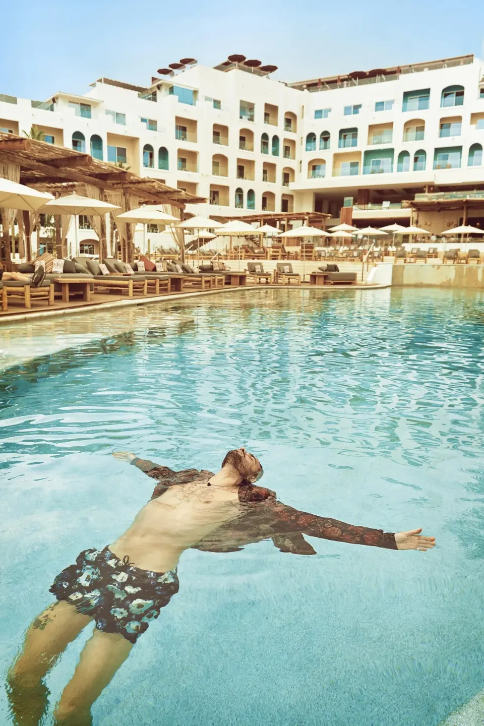 A guest floating in the resort pool with balconies behind him at ME Cabo.
