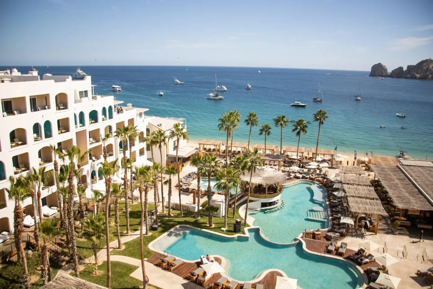 ME Cabo overlooks palm-lined pools, the beach, and boats in the bay.