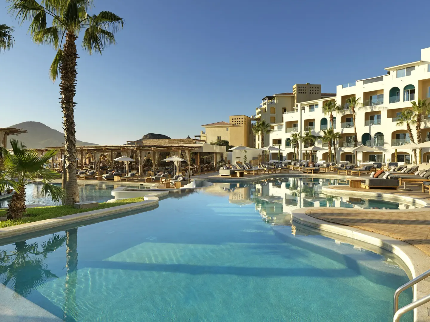 Resort pool with palm trees and white guest-room buildings at ME Cabo.