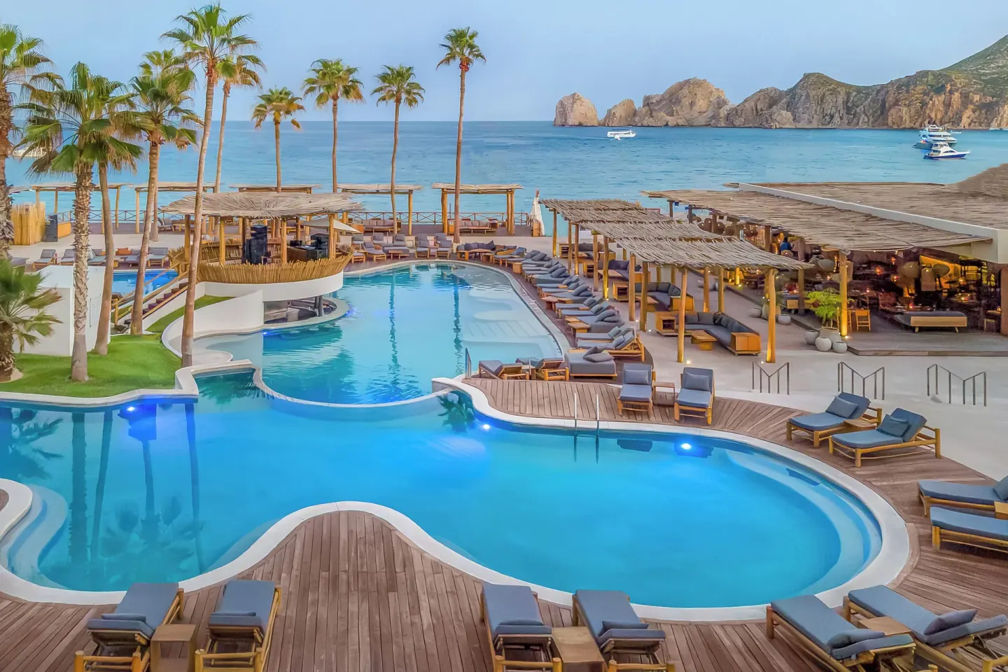 Curved resort pools, loungers, palm trees, and ocean views at ME Cabo.