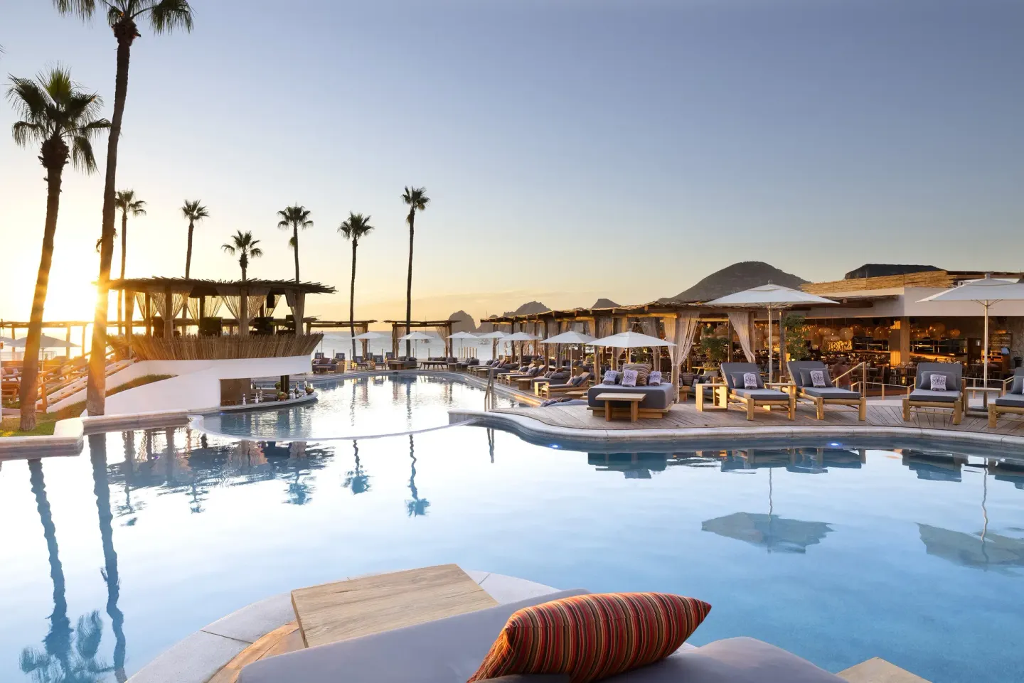 Sunset pool scene with palm trees, loungers, and open-air dining at ME Cabo.