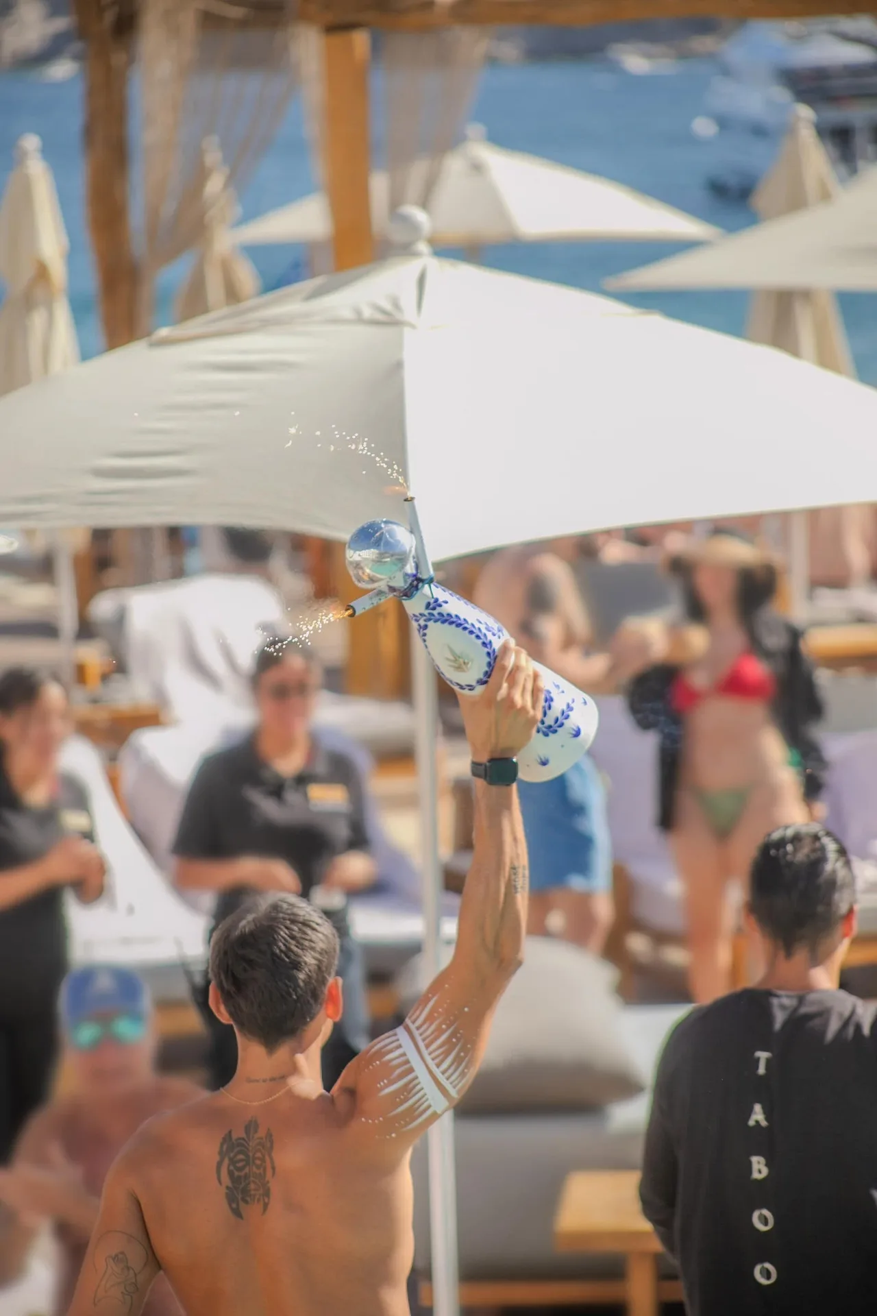 A beach club server sprays a large bottle among umbrellas and loungers at ME Cabo.