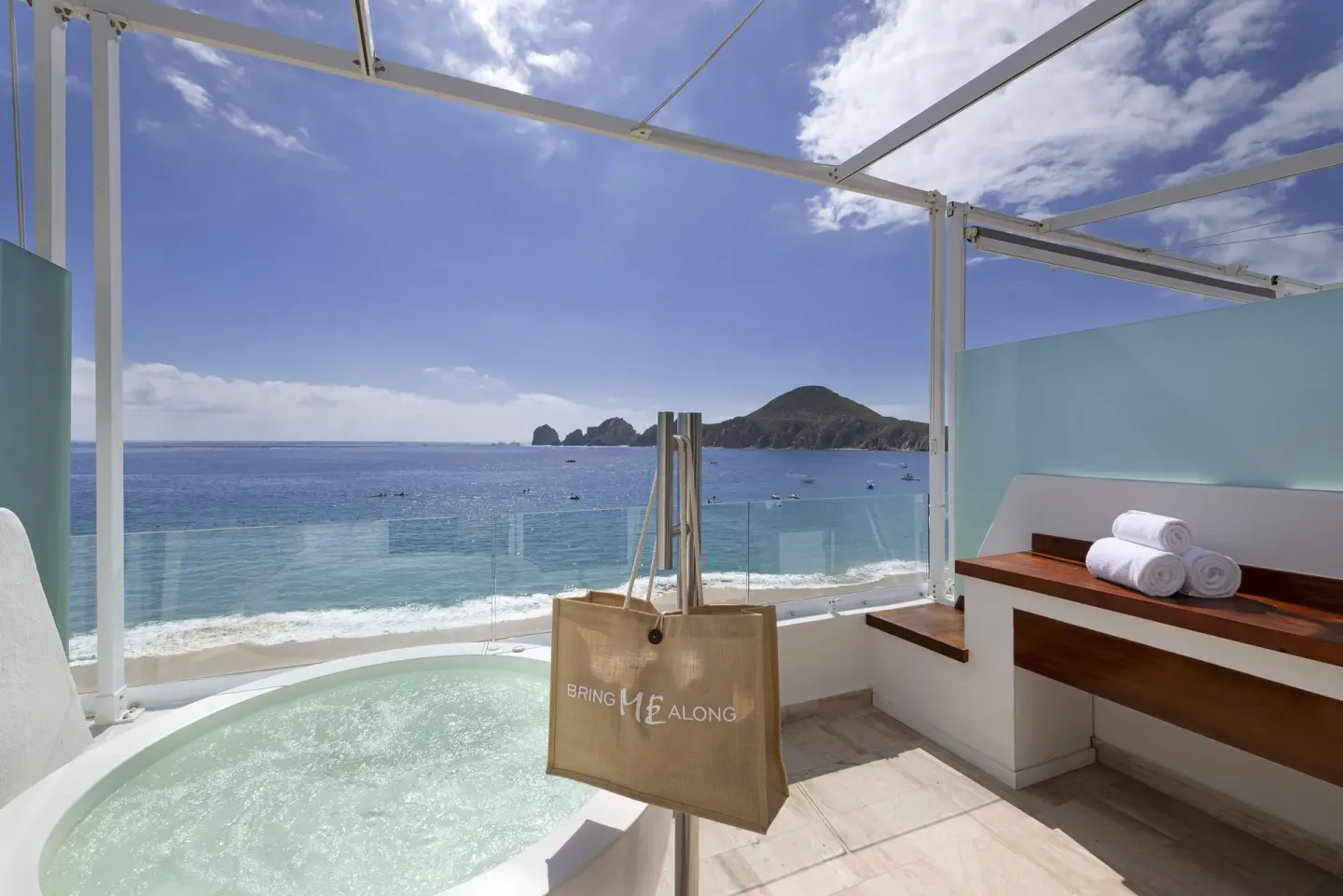 Hot tub terrace with towels, glass railing, beach and rocky ocean view at ME Cabo.