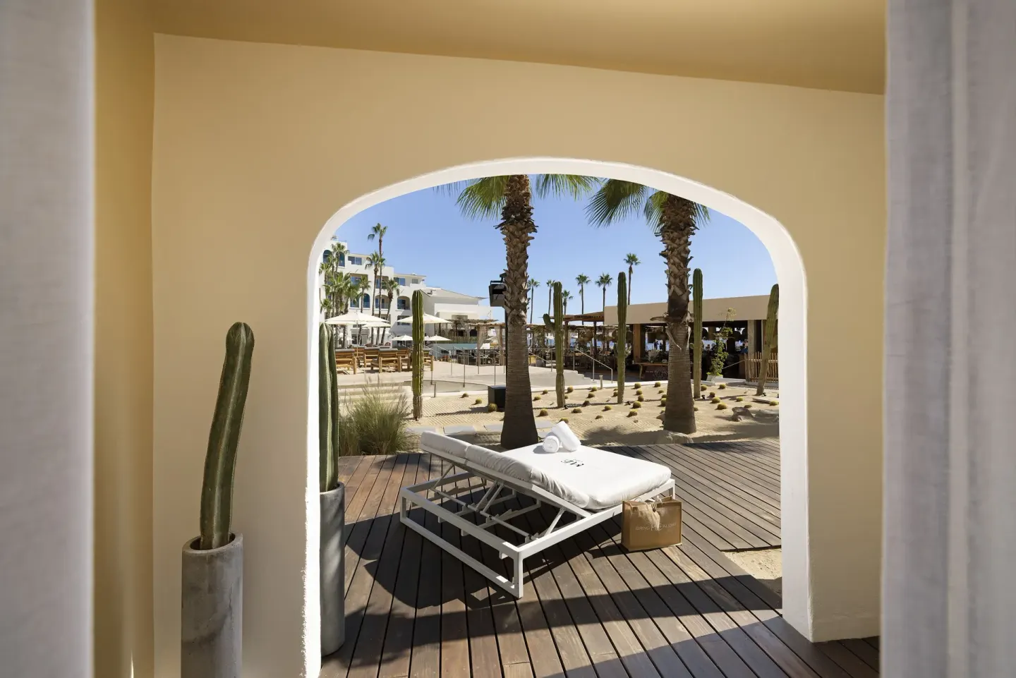 Superior ME+ terrace at ME Cabo viewed through an arch with a daybed, palms, cacti, and resort grounds.