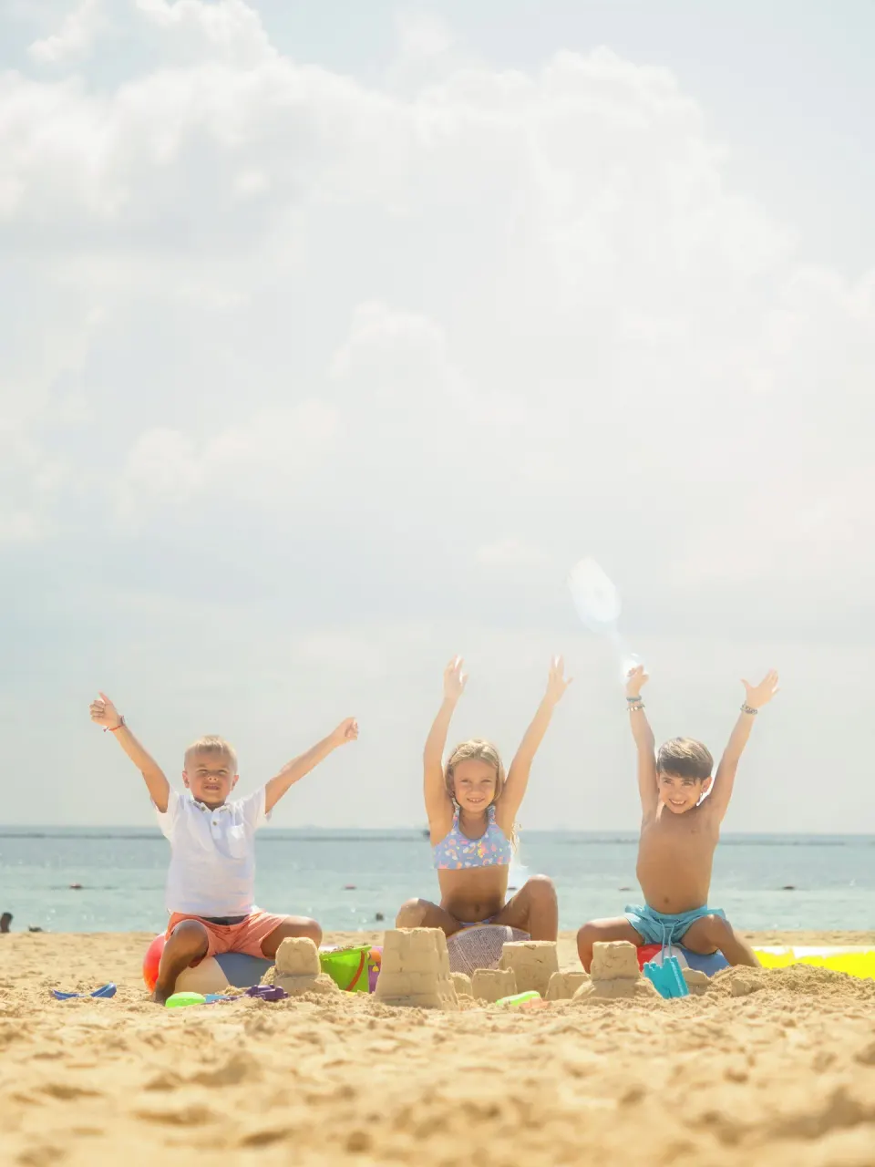 Three children sitting on the sand with arms raised beside their sandcastles on the beach at Melia Cozumel