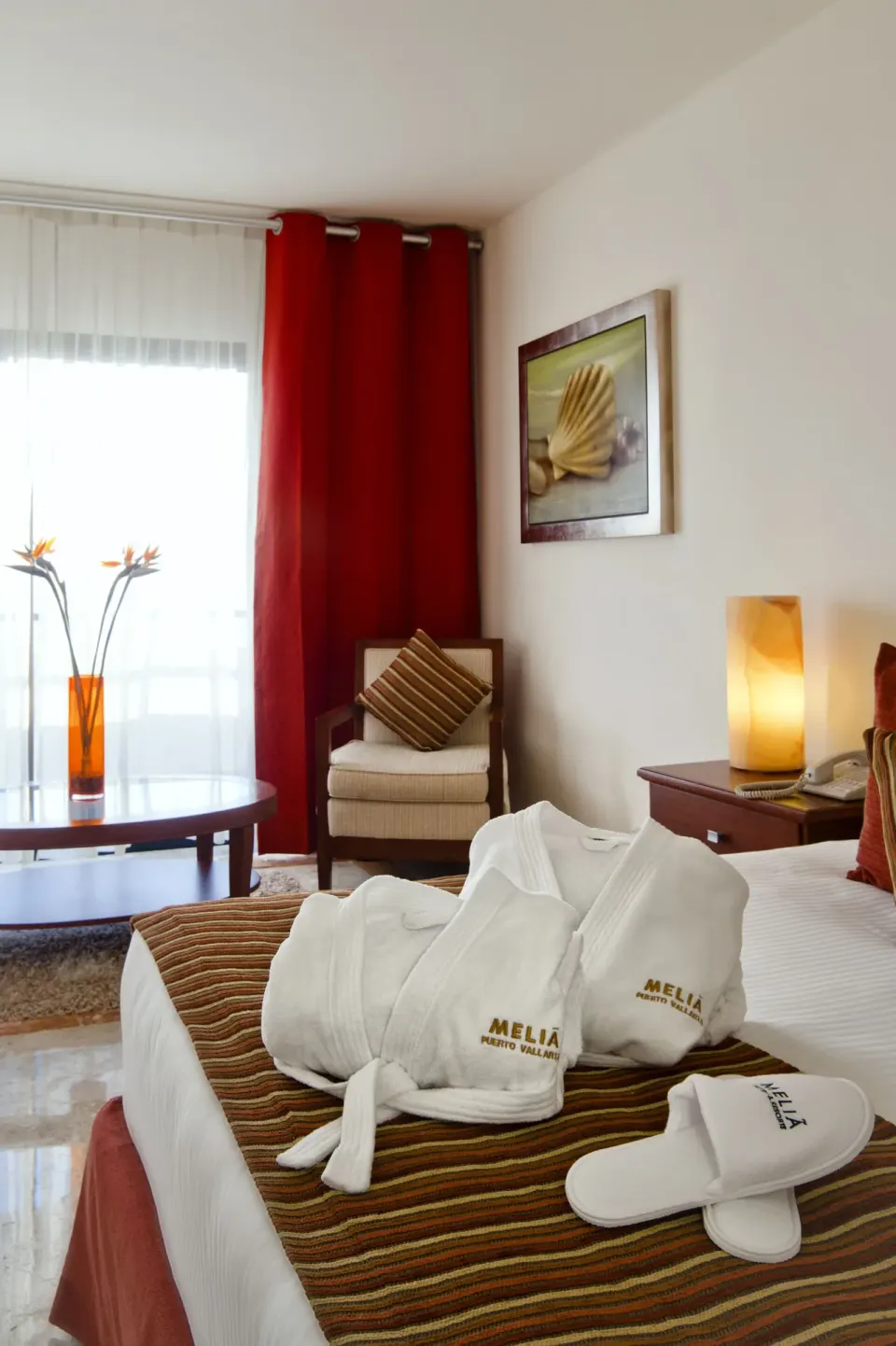 Guest room bed with robes, slippers, side chair, red curtains, and shell artwork at Melia Puerto Vallarta.